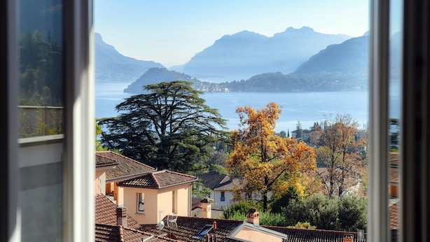 A view of a lake with mountains in the background, a tree with autumn leaves, and a building with a tiled roof.