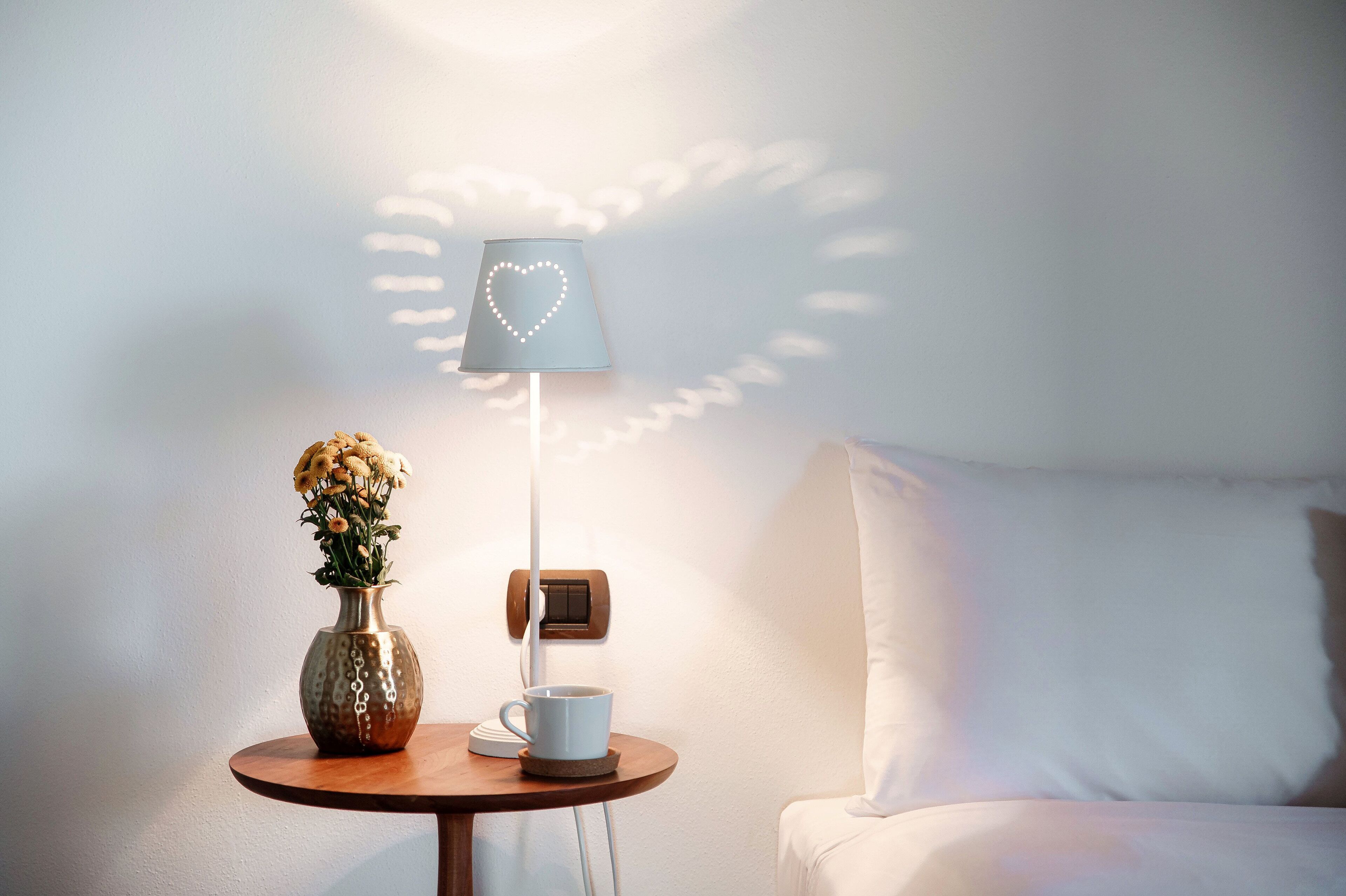 A bedside table with a vase of flowers, a cup, and a lamp with a heart-shaped light.