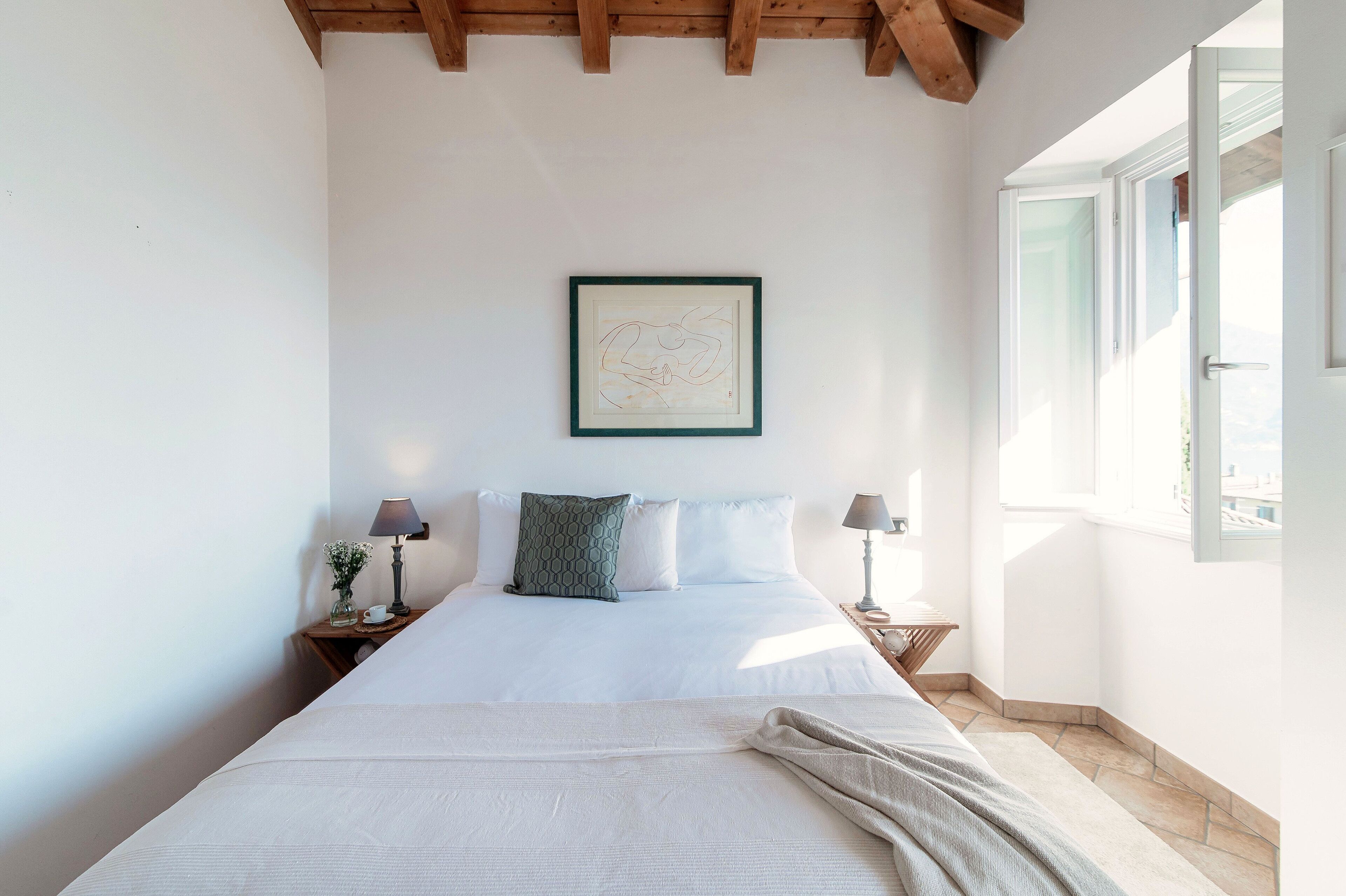 A neatly made bed with a green pillow, two bedside lamps, a framed artwork on the wall, and a window with a view.