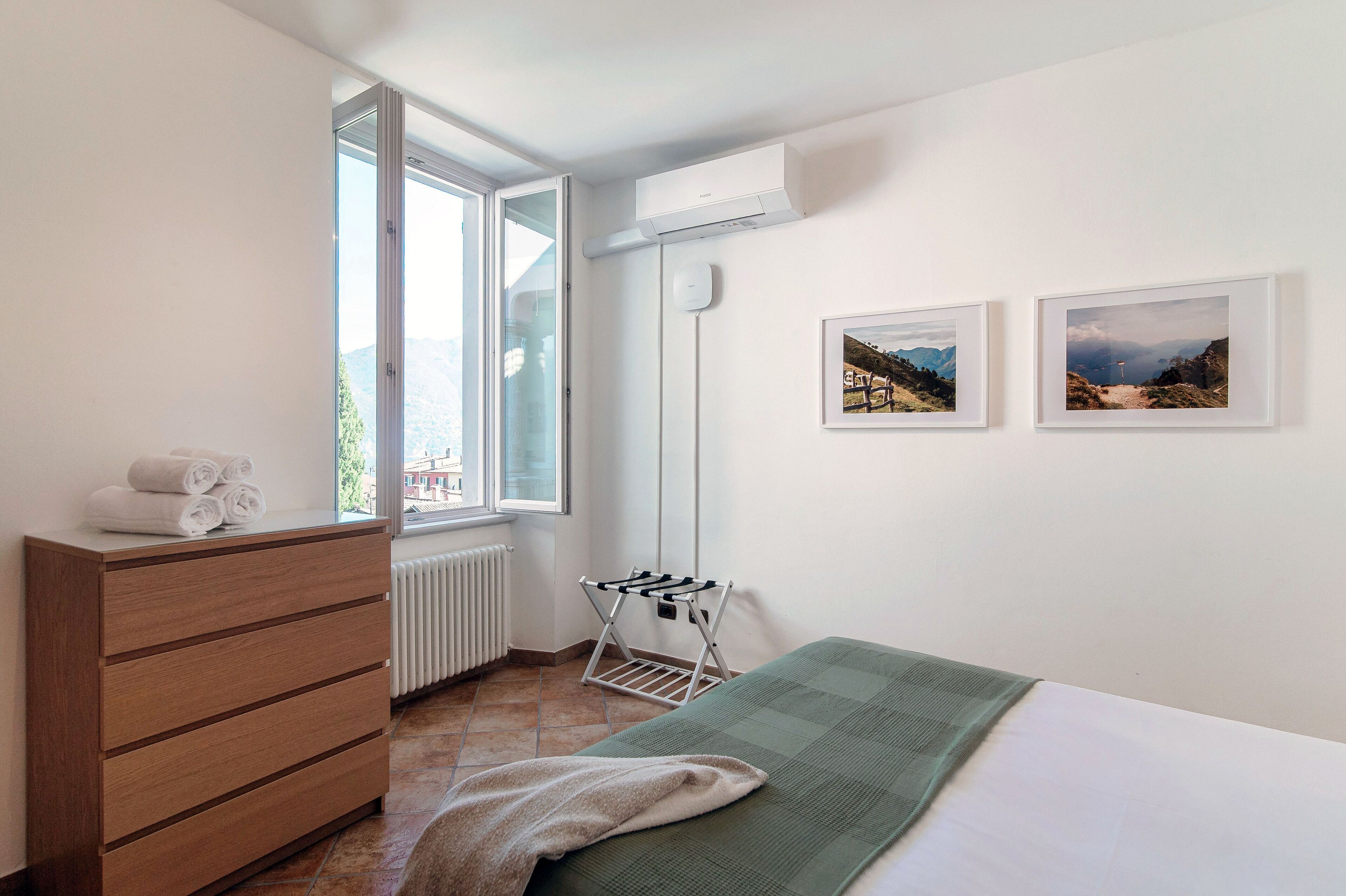 Een slaapkamer met een bed, een houten dressoir, een radiator, een raam en een wandgemonteerde airconditioner.