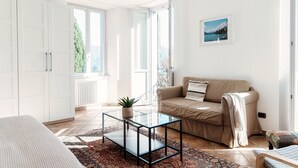 A bright living room with a glass coffee table, beige sofa, and a view of greenery through large windows.