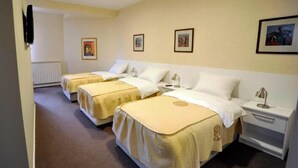 Camera d'albergo con tre letti singoli, ognuno dotato di una testiera bianca e una coperta beige. Sono presenti comodini con lampade e un piccolo tavolino con una lampada. La stanza ha un tappeto beige e pareti di colore chiaro.