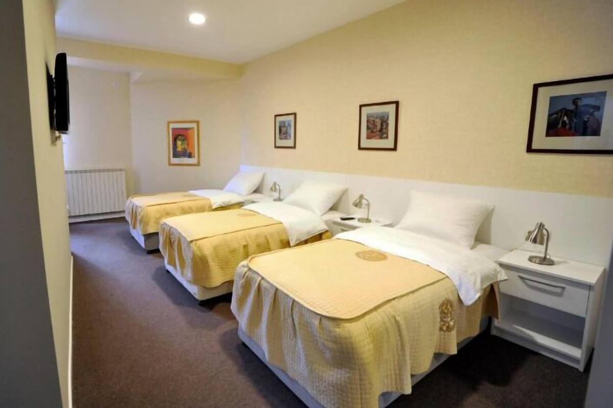 A hotel room with three single beds, each with a white headboard and a beige bedspread. There are bedside tables with lamps and a small bedside table with a lamp. The room has a beige carpet and light-colored walls.