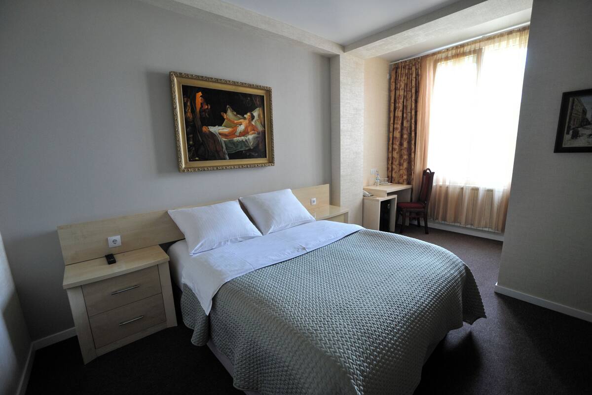A hotel room with a bed, a nightstand, a desk, a chair, and a window with curtains.