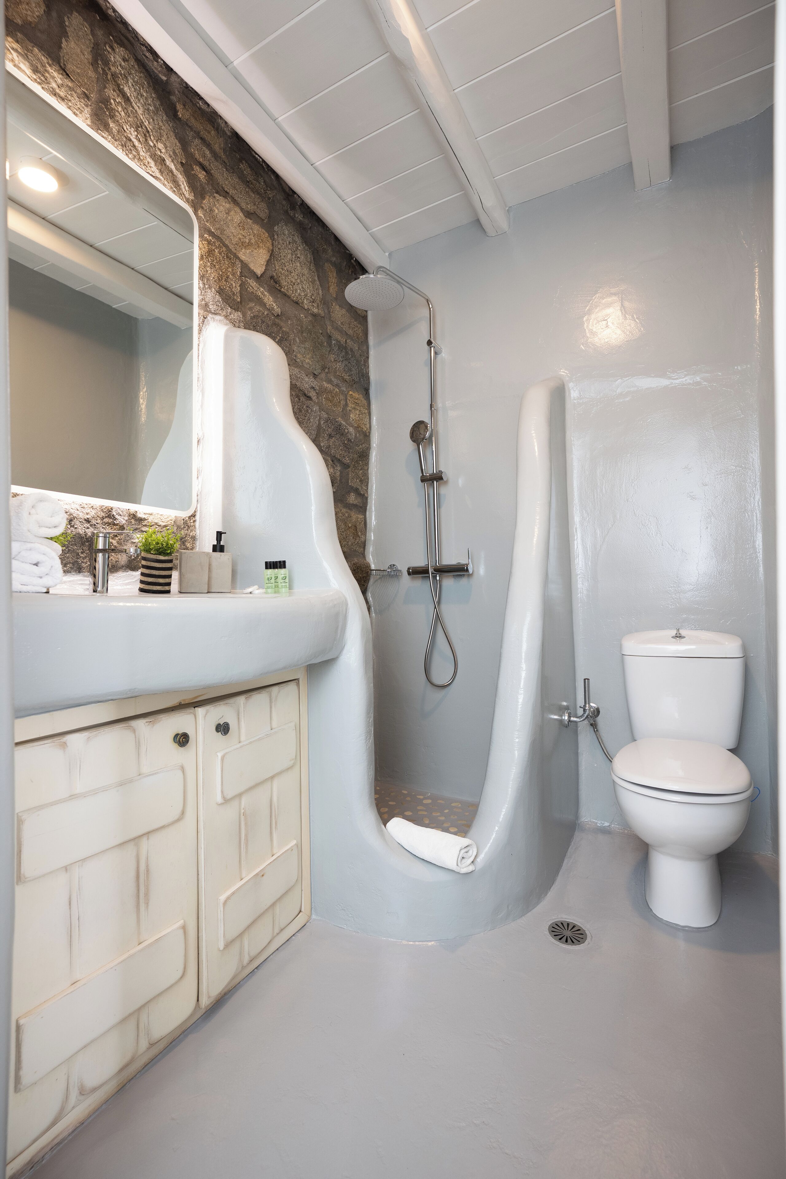 Een badkamer met een wit wastafelmeubel, een accentmuur van steen, een douche en een toilet.