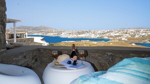 Une terrasse dotée de sièges blancs et confortables, une bouteille de vin et un verre posés sur une table, offrant une vue sur un paysage côtier avec des bâtiments et de l’eau.