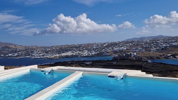 Une piscine à l’eau d’un bleu cristallin, entourée de structures blanches aménagées au bord du bassin, offrant une vue sur une ville côtière aux bâtiments blancs, ainsi que sur la mer et les collines lointaines.