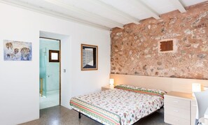 A bedroom with a stone wall, a bed with a floral bedspread, a bedside table with a lamp, and a door leading to another room.