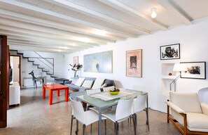 A modern living room with a glass dining table, white chairs, a red coffee table, a grey sofa, and a staircase leading to another level.