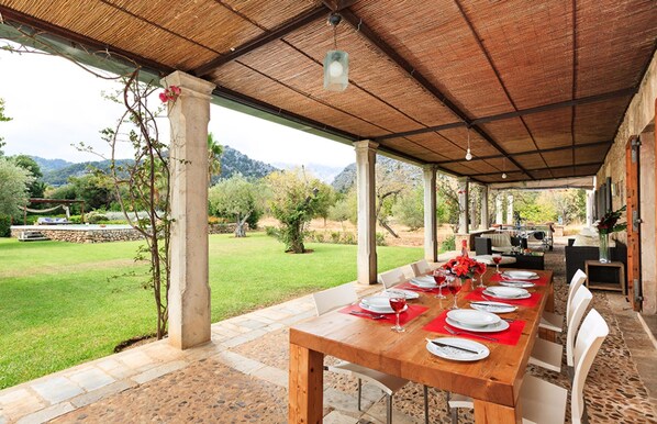An outdoor dining area with a long wooden table set for a meal, surrounded by stone pillars and a thatched roof.