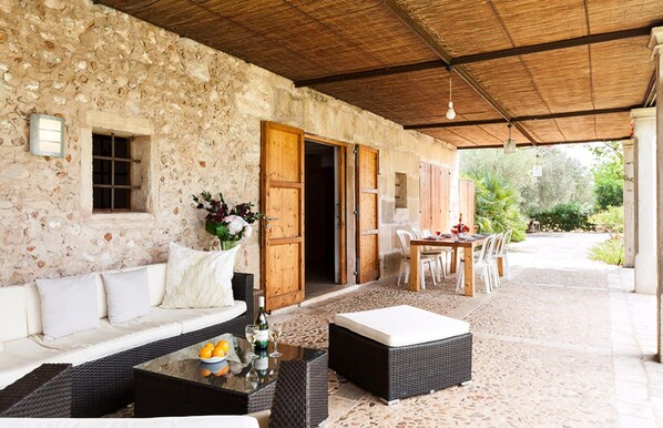 A covered outdoor seating area with a stone wall, a wooden door, a table with chairs, and a sofa.