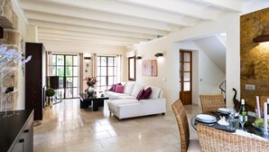 A spacious living room with a high ceiling, a white sofa, a dining area with wicker chairs, and a stone wall. - Elegant Villa with Stunning Views and Easy Beach Access (Pollenca)