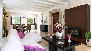 A spacious living room with a white sofa, a black coffee table, and a fireplace. There is a dining area in the background with a table and chairs. - Elegant Villa with Stunning Views and Easy Beach Access (Pollenca)