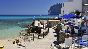 A coastal area with a clear view of the sea, a rocky shoreline, and a building with outdoor seating. - Elegant Villa with Stunning Views and Easy Beach Access (Pollenca)