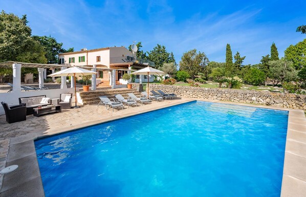 A poolside area with lounge chairs and umbrellas, a stone wall, and a building with a white facade and green shutters.