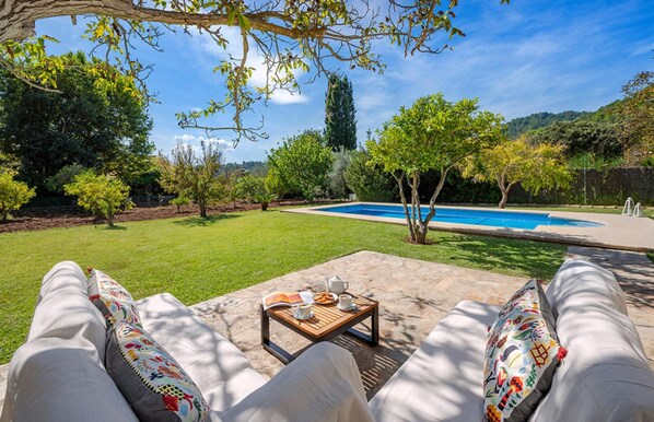 A poolside area with a wooden table, two lounge chairs, and a view of a swimming pool surrounded by trees and greenery.