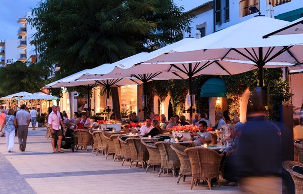 An outdoor café with wicker tables and chairs, covered by large umbrellas, and people dining.