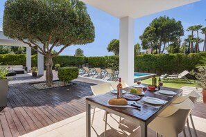 Un espace extérieur aménagé pour les repas, avec une table dressée pour deux personnes, entouré de chaises blanches, d’une terrasse en bois et, au fond, d’une piscine.