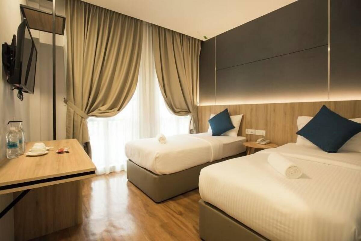 Ein Hotelzimmer mit zwei Betten, einem Holztisch, einem Flachbildfernseher und einem großen Fenster mit Vorhängen.