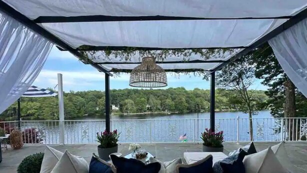 Un área con mesas y sillas al aire libre, cubierta por un toldo blanco, con una lámpara colgante y vistas a un lago y árboles.