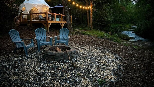 En rustik utomhusplats med sittplatser, Adirondack-stolar och en eldstad, omgiven av en skog och ett tält.