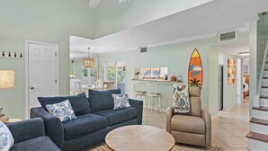 A spacious living area with a sofa, armchair, and round coffee table. A surfboard decoration is visible on the wall. - Seascape 55b- Surfer Girl (Miramar Beach)