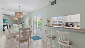 A dining area with a glass table and white chairs, a kitchen with white cabinets, and a view through the glass doors. - Seascape 55b- Surfer Girl (Miramar Beach)