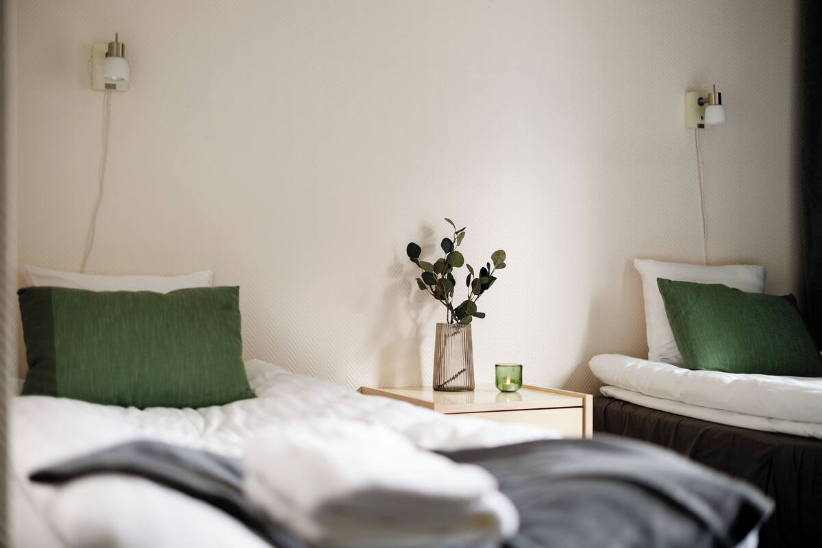 A neatly made bed with white and green bedding, a small bedside table with a green candle, and a vase with green leaves.