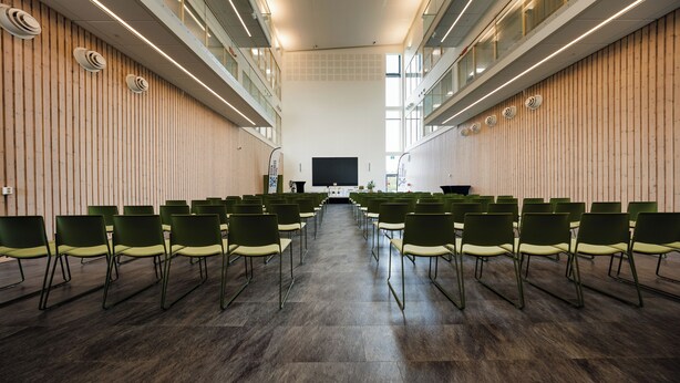 A conference room with rows of chairs facing a large screen, wooden panel walls, and a high ceiling with linear lighting. - Hotell Säsong - Boden Business Park (Boden)