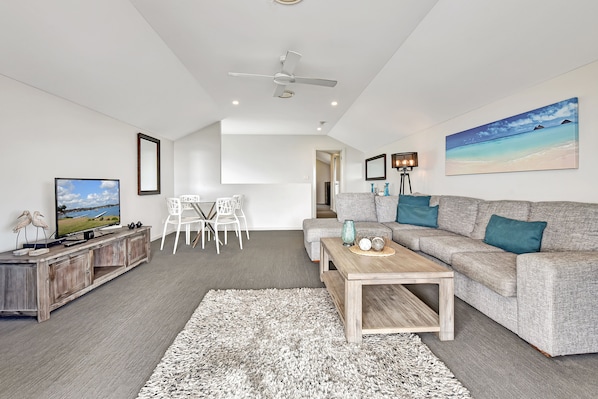 Un salon moderne avec un canapé gris, une table basse, un téléviseur et un tableau représentant une scène de plage.