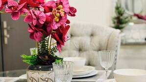 Una mesa de comedor dispuesta con platos, tazones y un vaso blancos. Un florero con flores rosas está sobre la mesa.