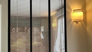 Een kamer met een groot raam met zonwering, een wandlamp en een wit kussen op een wit hoofdeinde.
