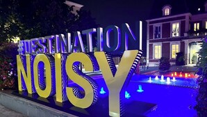 Een groot verlicht bord met de tekst 'DESTINATION NOISY', met een fontein en een gebouw op de achtergrond.