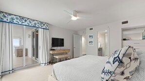 A modern bedroom with a large bed, a flat-screen TV, a ceiling fan, and a sliding glass door leading to a bathroom. - Seaspray West 801- Gulf Front (Pensacola)