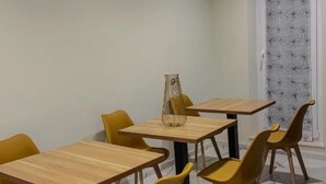 Un coin repas avec des tables en bois et des chaises jaunes, un vase posé sur l’une des tables, et un rideau à motifs en arrière-plan.