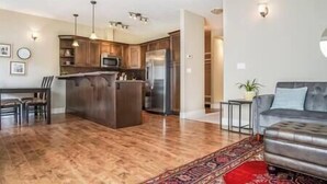 A modern kitchen with wooden cabinets, a central island, and stainless steel appliances. There is a dining area with chairs and a sofa in the living room.