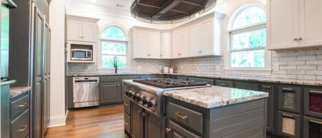 A modern kitchen with dark cabinetry, a central island, stainless steel appliances, and large windows.