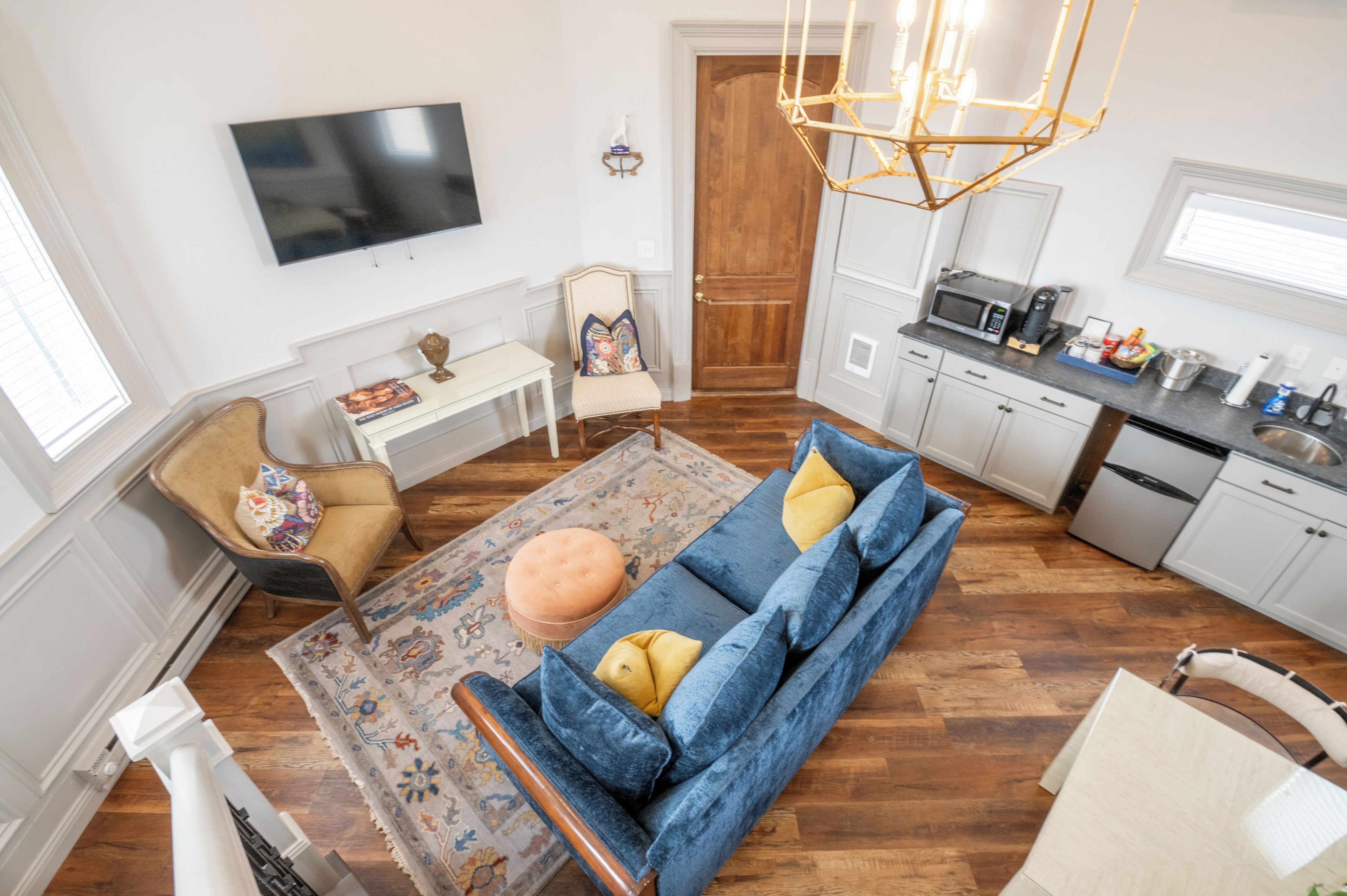Un soggiorno moderno con un divano blu, un pavimento in legno, una TV a muro e un angolo cucina con un forno a microonde e pensili.