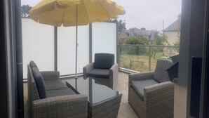 Varanda com móveis de vime, uma mesa de vidro e um guarda-sol amarelo.