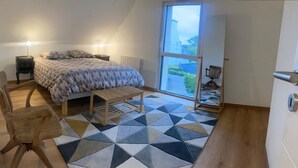 Quarto com cama, mesas de cabeceira, uma cadeira, um banco e uma janela grande.