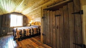 Une pièce rustique dotée de planchers en bois et d’un plafond en pente, comprenant un lit recouvert d’une couverture à motifs, une grande armoire en bois et une fenêtre laissant entrer la lumière naturelle.