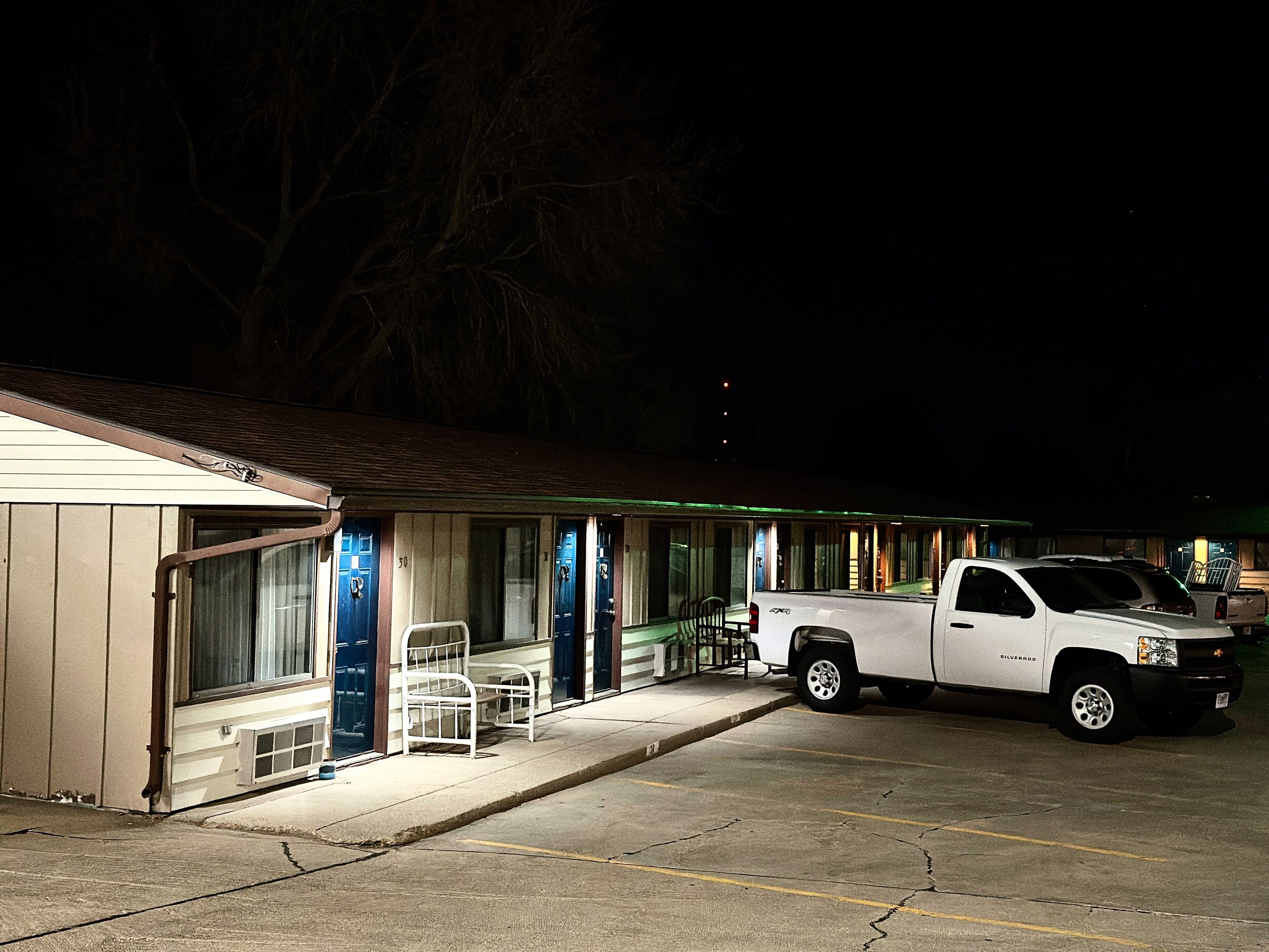 A motel with a white pickup truck parked in the driveway, a covered outdoor seating area, and a row of doors with numbers.