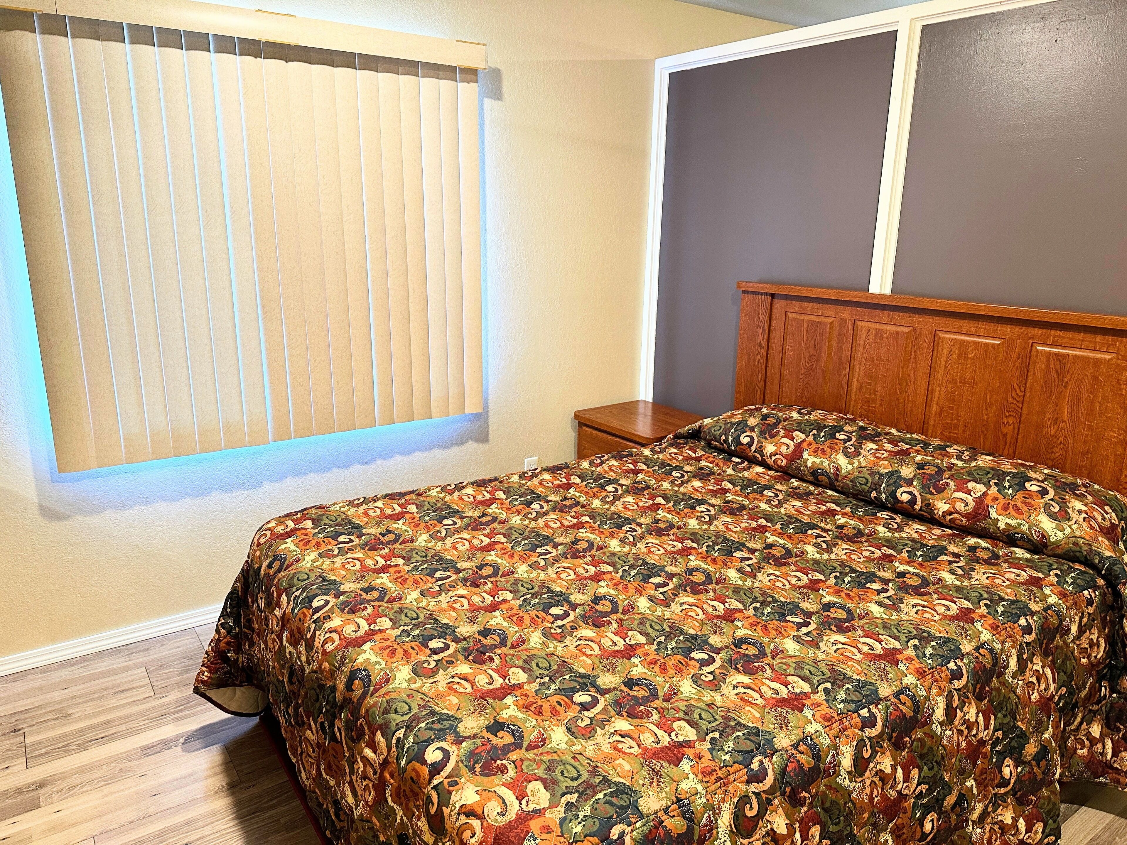 A bedroom with a bed, a nightstand, a window with blinds, and a wooden headboard.