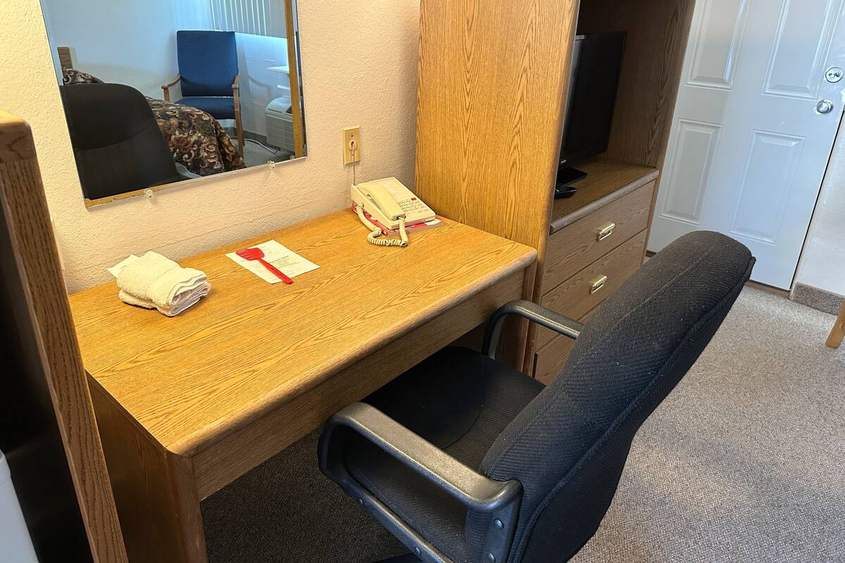 A compact hotel room with a wooden desk, a chair, a small vanity with a mirror, a telephone, and a television.