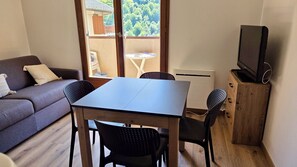 A room with a sofa, a table, two chairs, a television on a wooden cabinet, and a view of trees through a window. - Apartment in Valloire at the foot of the slopes (Valloire)