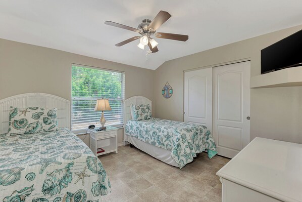 A bedroom with two beds, a ceiling fan, a window with blinds, a bedside table with a lamp, and a wall-mounted TV.
