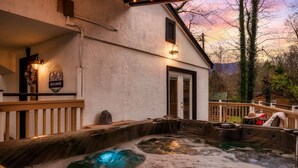 A spa area with a hot tub, a wooden deck, and a building with a sign that reads "OUTLANDER". - Outlander Couples Cottage/Walk to Downtown/Hot Tub (Gatlinburg)