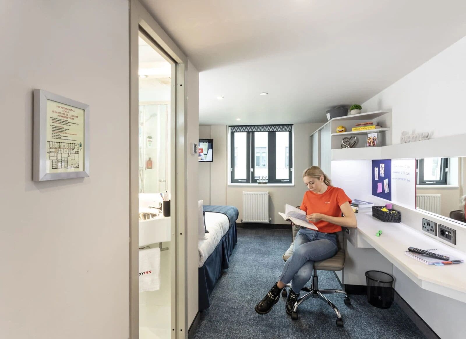 Un dormitorio pequeño y moderno con una cama, un escritorio y una silla. Hay una ventana con vistas al exterior, un radiador y un televisor montado en la pared.