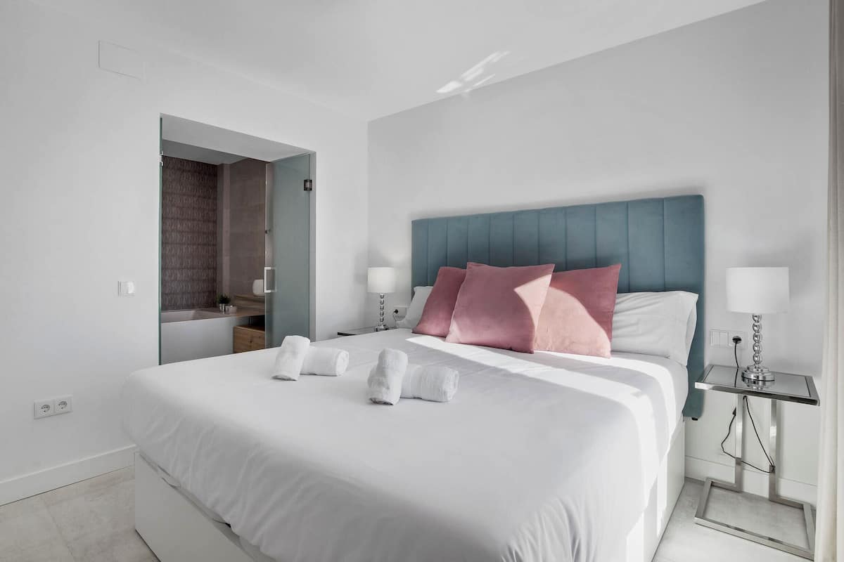 A modern hotel room with a large bed, a nightstand with lamps, and a bathroom visible through an open door.