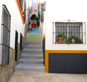 A narrow alleyway with a staircase leading to a building. There are potted plants on the steps and a window with a metal grill on the building.